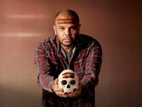 Man looks into camera holding a skull. He and the skull have brown lines painted across the forehead