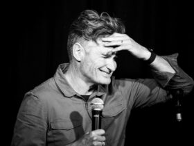 Dave holds a microphone. His hand is on his forehead and he is smiling.