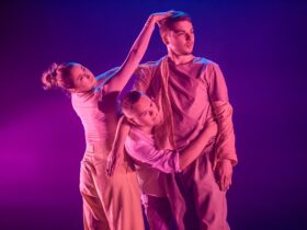 Three dancers stand together. Two of them embrace. The third has her hand on the head of another.