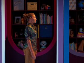 A blonde woman stands side-on to camera. Beside her a bookshelf filled with toys, books & photos