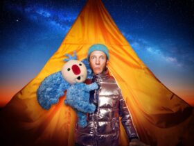 A man, holding a big-eared blue koala, emerges from an orange tent and lots disconcertingly at us.