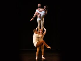 Woman stands. A man stands on her shoulders. Another man hangs off her neck. First man holds a woman