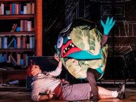 A person in a scary lettuce costume stands over a man lying on the floor dressed as a school boy.