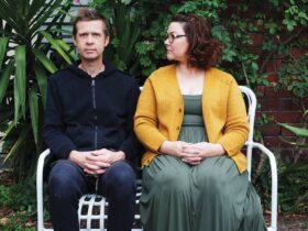 Two artists sitting down, one is looking at the camera and one is looking at the other.