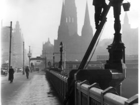Black and while photo- a man wears a suit and hat has climbed up a ladder and is cleaning a lamppost