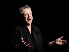 A middle aged short-haired woman sings happily in darkness while holds her hands in an open gesture.