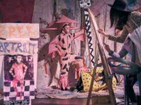Man in a pink costume with a large hat poses in front of three people drawing him.