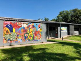 Photograph of grey building with colourful mosaic, surrounded by green lawns and trees.
