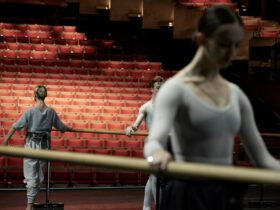Three dancers holding the barre on stage. One is facing towards the camera and two facing away.