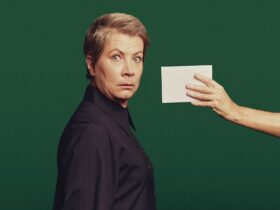 A short-haired woman looks at camera as though startled. An outstretched arm offers her an envelope